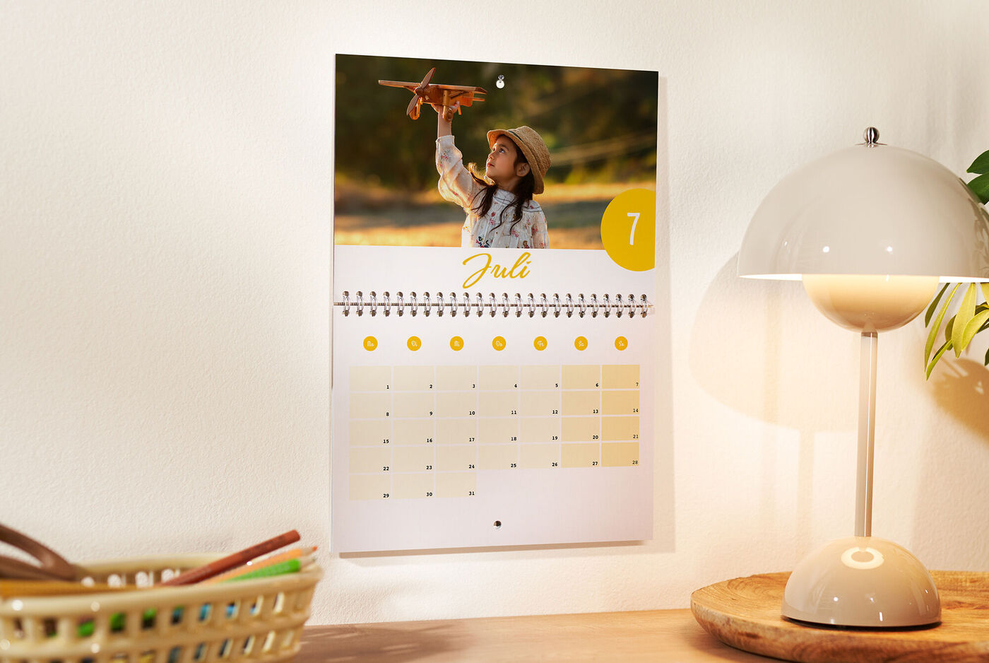 Ein Wandkalender zeigt ein fröhliches Kind, das ein Spielzeugflugzeug hält. Der Monat Juli ist hervorgehoben.